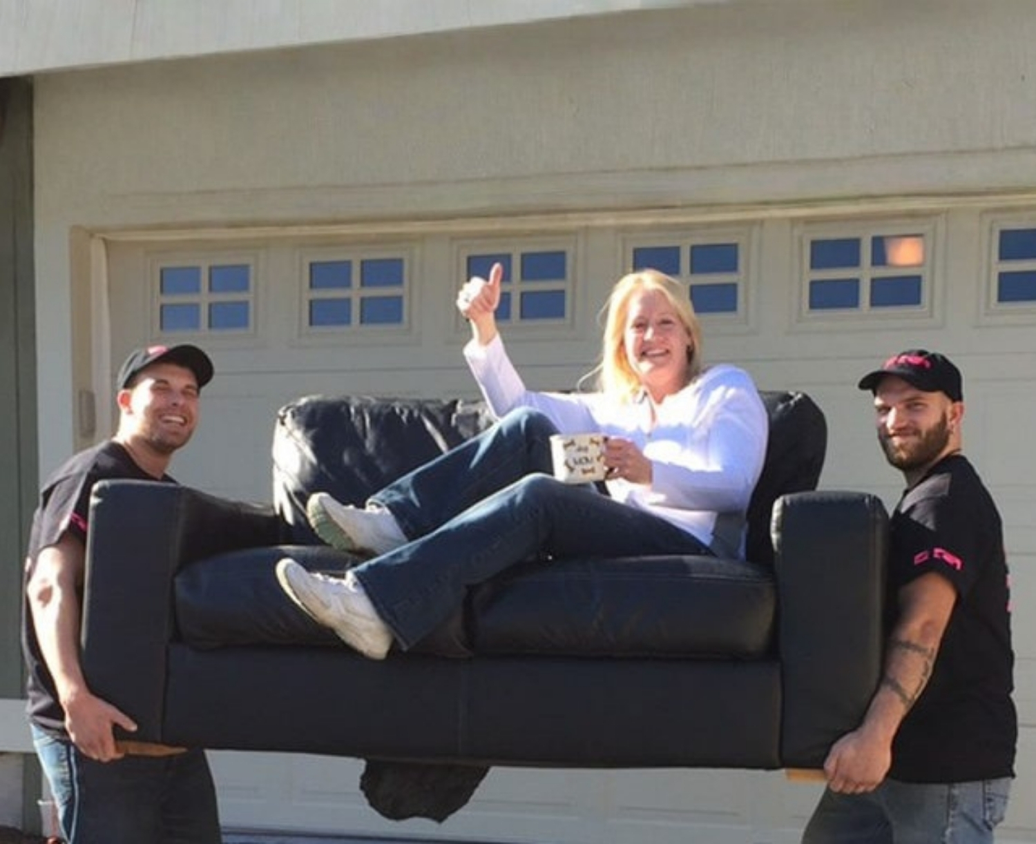 Two movers carry a sofa with a smiling homeowner seated on it, illustrating whether to move or replace furniture.